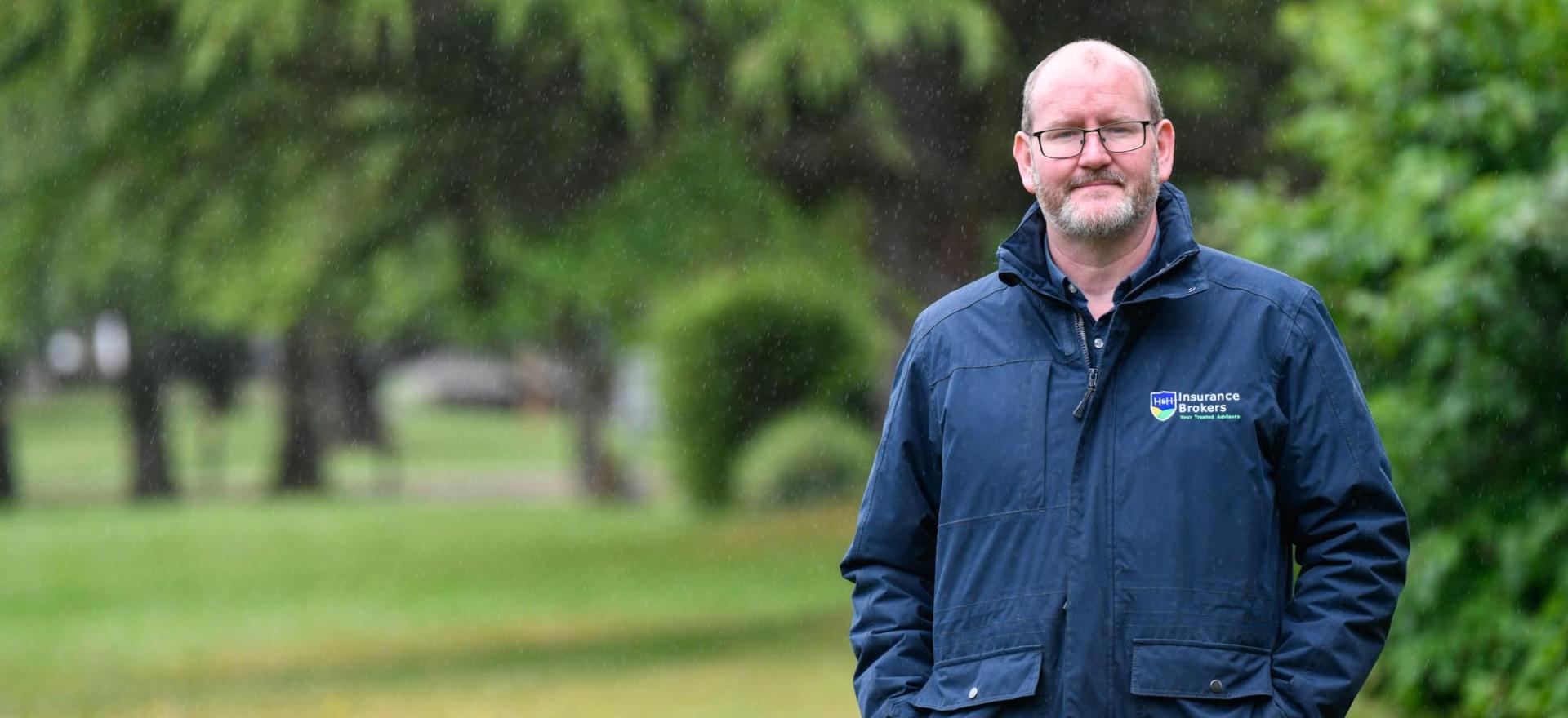 Stuart Torrance, Claims Manager at H&H Insurance Brokers standing in the rain, with a background of grass and hedges