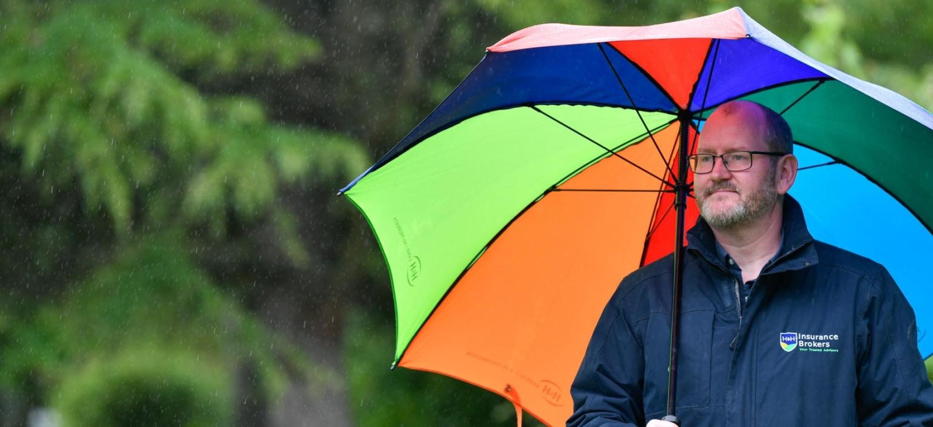 Stuart Torrance, Claims Manager at H&H Insurance Brokers standing in the rain under a colourful umbrella